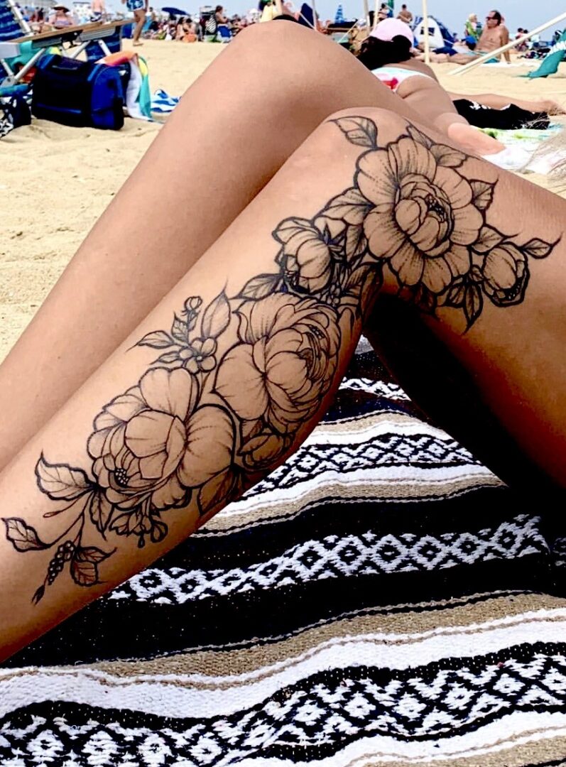 A woman with tattoos on her legs sitting in the sand.