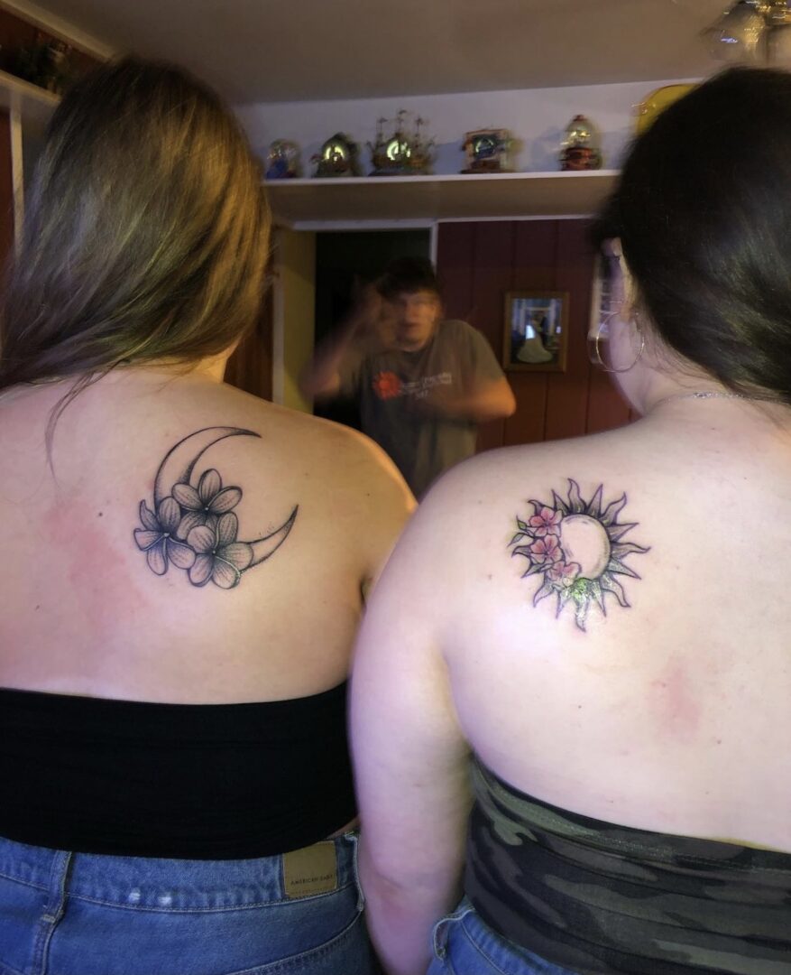 Two women with tattoos on their backs looking at a mirror.