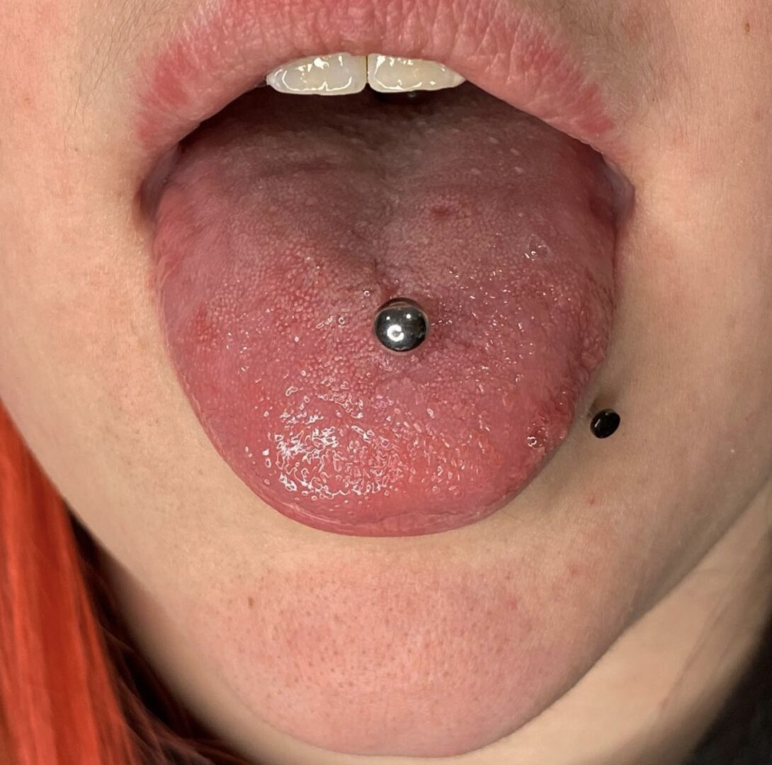 A woman with red hair and black nails piercing her tongue.